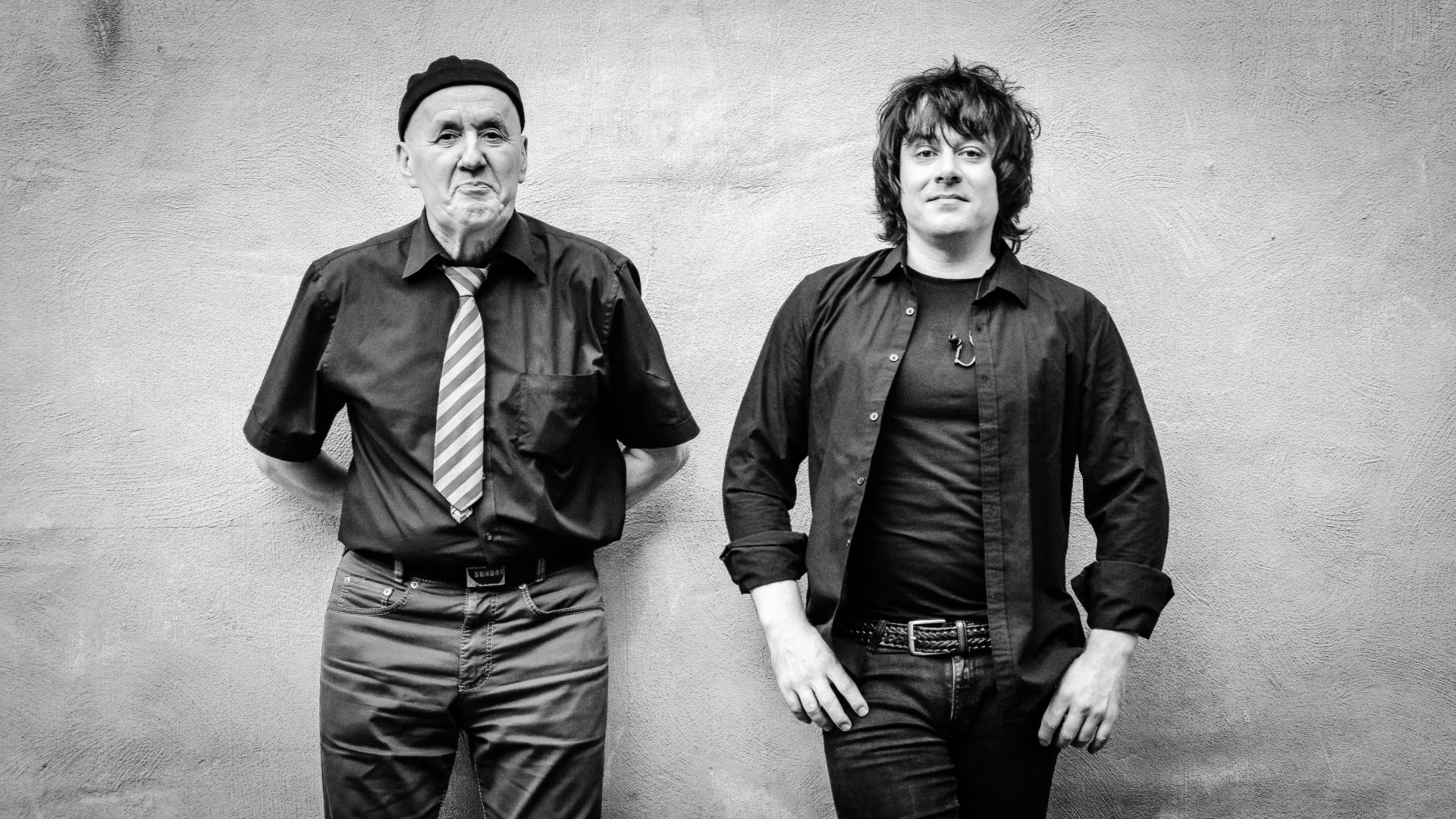 A black and white portrait of two musicians Thommi and Leon standing side by side against a textured wall, one older man wearing a black shirt and a hat, and a younger man with tousled hair dressed in a black shirt.